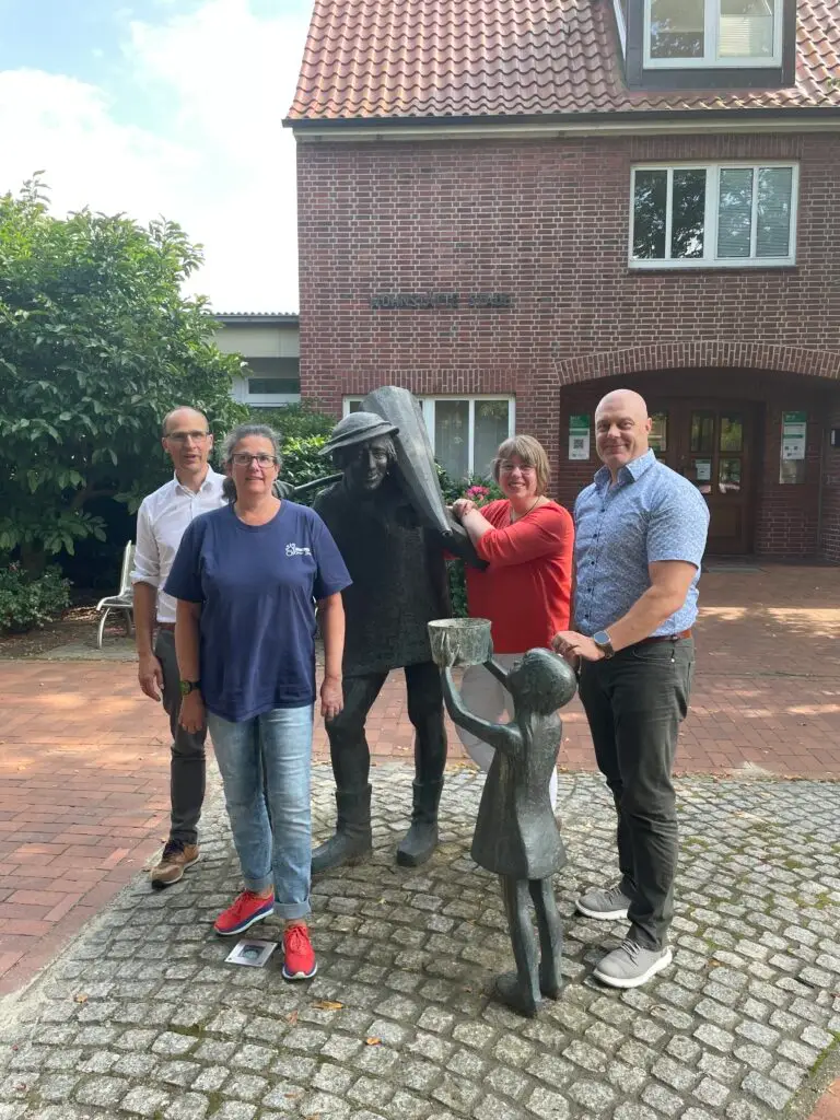 Vier Erwachsene stehen im Freien neben zwei Bronzestatuen - einer großen Figur und einem Kind, das einen Eimer hält - auf einem gepflasterten Platz vor einem roten Backsteingebäude mit Grün im Hintergrund.