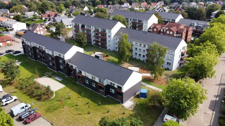 Luftaufnahme eines Wohnviertels mit modernen Wohngebäuden, parkenden Autos, grünen Rasenflächen, Bäumen und umliegenden Häusern an einem sonnigen Tag.