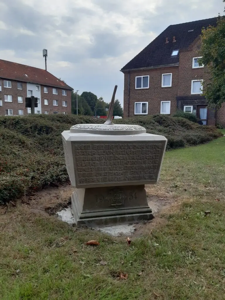 Eine steinerne Sonnenuhr mit eingraviertem Text steht auf einer Wiese vor einem Wohnhaus aus Backstein. Der Himmel ist bewölkt, und hinter der Sonnenuhr sind Büsche zu sehen.