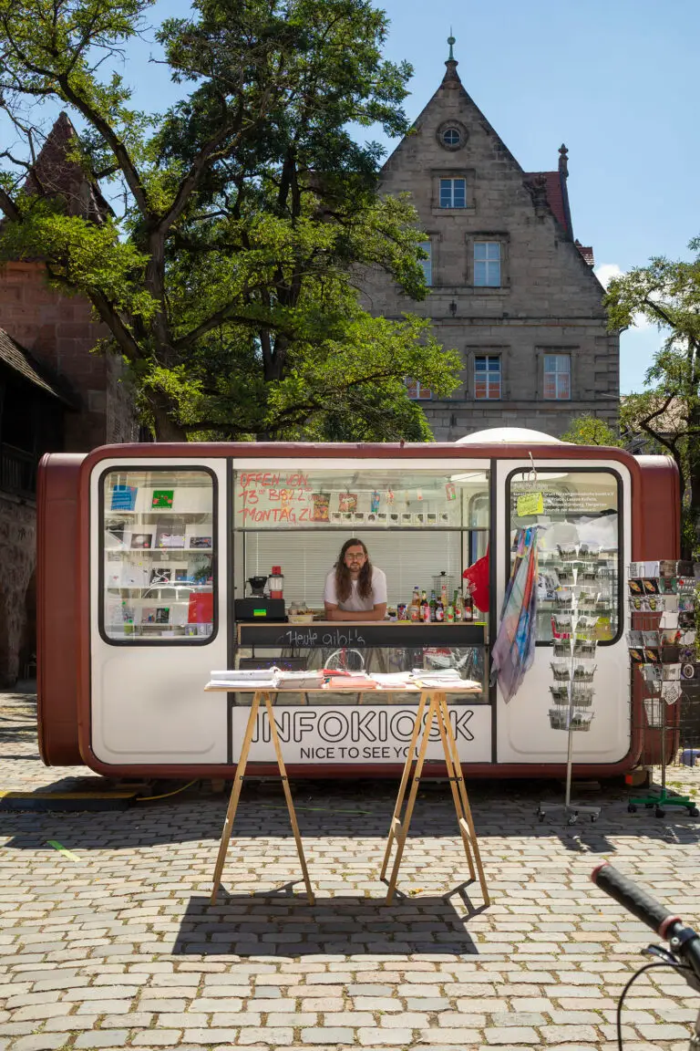 Ein kleiner Infokiosk mit großen Fenstern steht an einer Kopfsteinpflasterstraße. Eine Frau steht darin, umgeben von Broschüren, Postkarten und Souvenirs. Im Hintergrund sind ein hohes historisches Gebäude und Bäume zu sehen.
