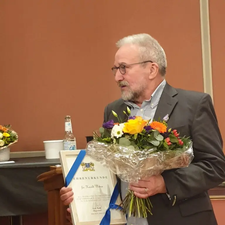Ein älterer Mann im Anzug hält einen Strauß bunter Blumen und eine gerahmte Urkunde in der Hand. Er steht drinnen neben einem Tisch mit einer Flasche und anderen Blumen, schaut nach rechts und lächelt.