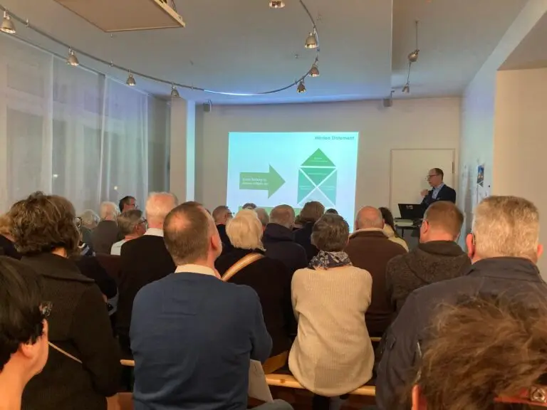 Eine Gruppe von Personen sitzt vor einer Leinwand, auf die eine Präsentation projiziert wird. Ein Redner steht auf einem Podium und spricht zu den Zuhörern. Der Raum ist modern beleuchtet und verfügt über große Fenster. Die Folie zeigt ein Diagramm mit Text.