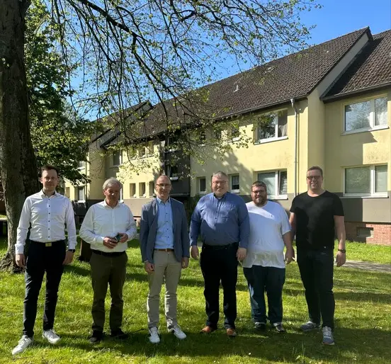Sechs Männer stehen an einem sonnigen Tag auf einer Wiese vor einem Wohnhaus, wobei die Bäume Schatten werfen und grüne Blätter über ihnen zu sehen sind.