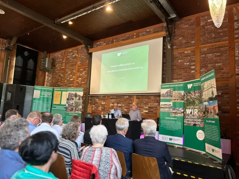 Eine Sitzung findet in einer gemauerten Halle statt, in der die Teilnehmer gegenüber einer Bühne sitzen. Zwei Redner sitzen an einem Tisch mit Mikrofonen. An der Vorderseite des Saals sind Informationsbanner und eine Präsentationswand zu sehen.