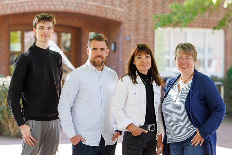 Vier Personen stehen im Freien vor einem Backsteingeb&auml;ude und l&auml;cheln in die Kamera. Es scheint sich um zwei M&auml;nner und zwei Frauen zu handeln, die leger gekleidet sind, mit Gr&uuml;n und Sonnenlicht im Hintergrund.