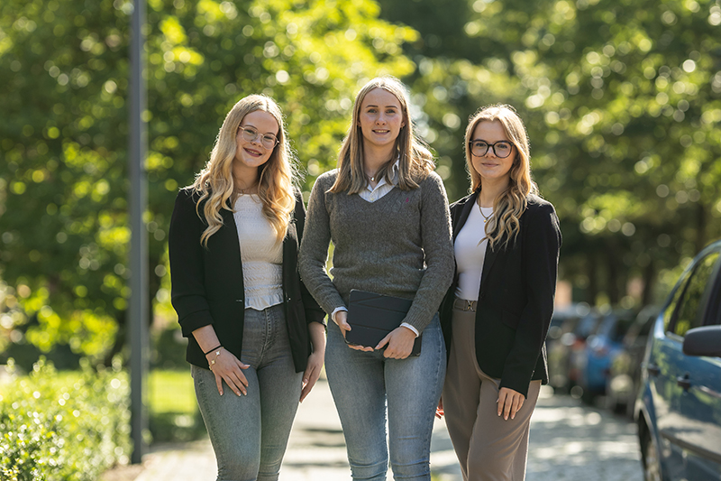 Drei junge Frauen stehen an einem sonnigen Tag Seite an Seite im Freien und l&auml;cheln in die Kamera. Sie sind leger gekleidet, im Hintergrund sind B&auml;ume und geparkte Autos zu sehen.