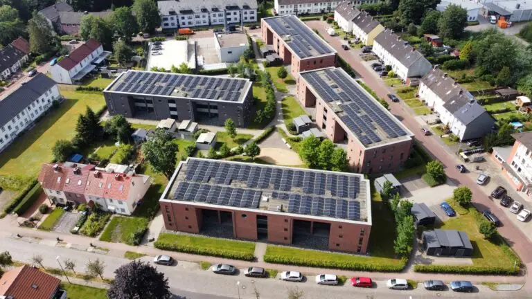 Luftaufnahme von Wohngebäuden mit Sonnenkollektoren auf den Dächern, umgeben von Bäumen, parkenden Autos und Grünflächen in einem Vorort.