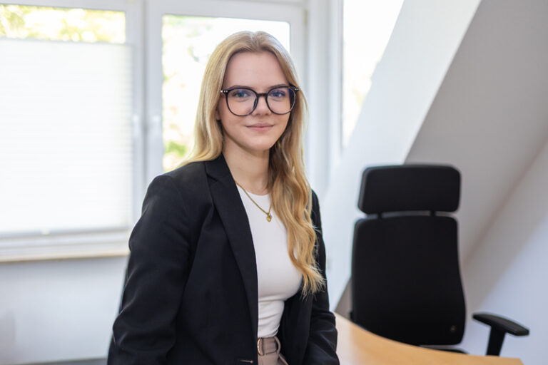 Véronique Denker. Sie ist Auszubildende der Wohnstätte Stade eG. Frau Denker ist früheren Alters, hat hüftlange blonde Haare und trägt ein schwarzes Jackett und ein weißes Hemd.