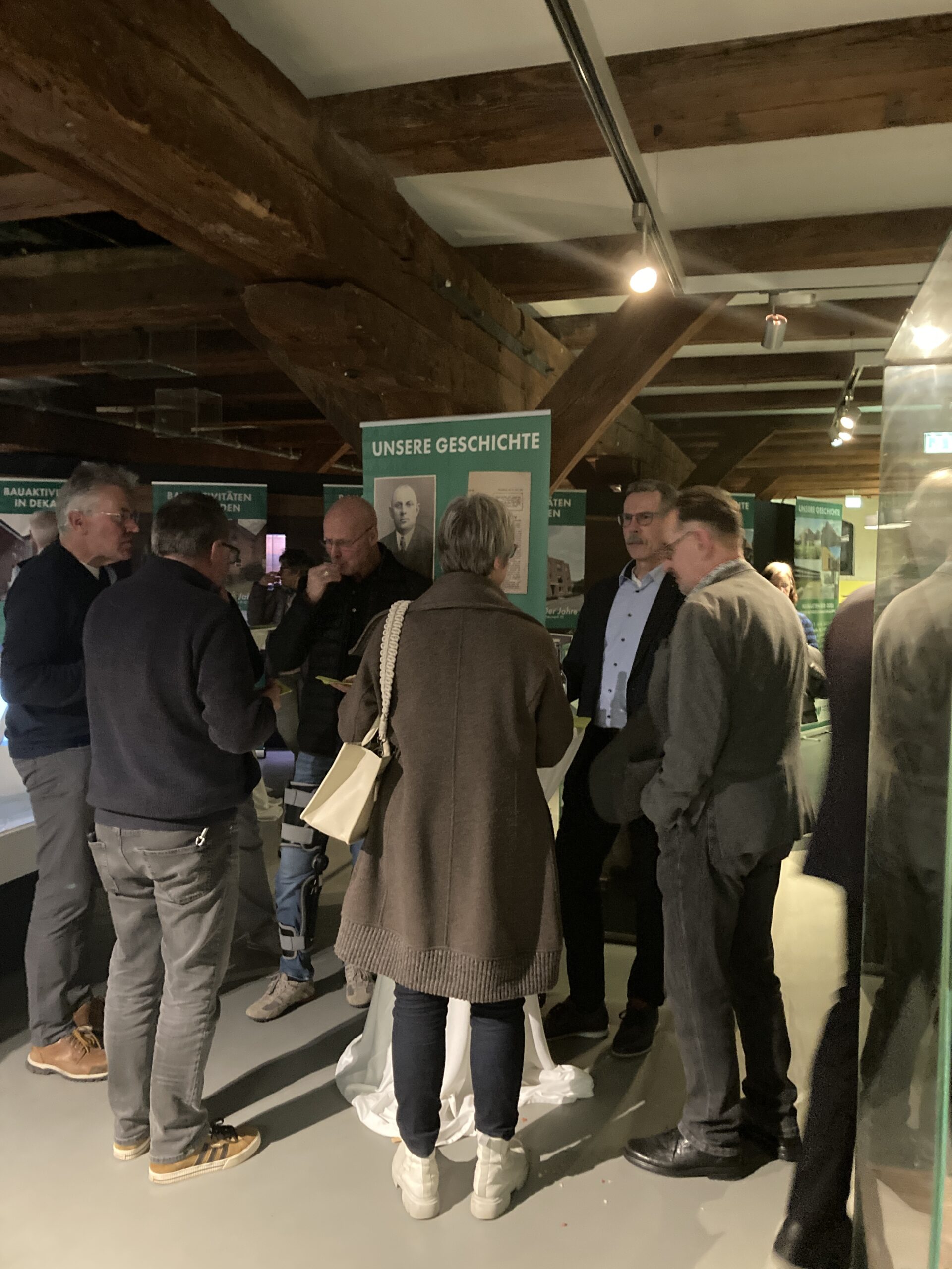 Eine Gruppe von Menschen steht und unterh&auml;lt sich in einem &uuml;berdachten Ausstellungsraum mit Holzbalken. Hinter ihnen steht ein gr&uuml;nes Schild mit der Aufschrift UNSERE GESCHICHTE auf Deutsch. Die Beleuchtung ist warm und konzentriert.