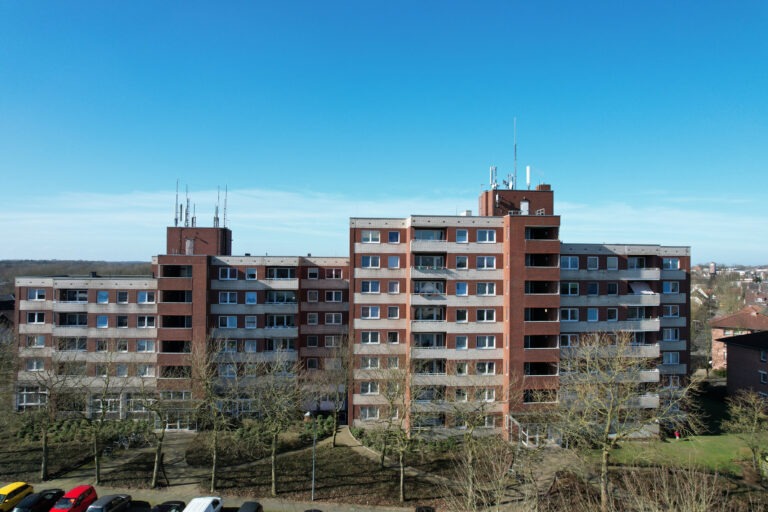 Ein mittelhoher roter Backsteinwohnungsbau mit Balkonen und Antennen auf dem Dach vor einem klaren blauen Himmel. Im Vordergrund sind blattlose Bäume und geparkte Autos zu sehen.