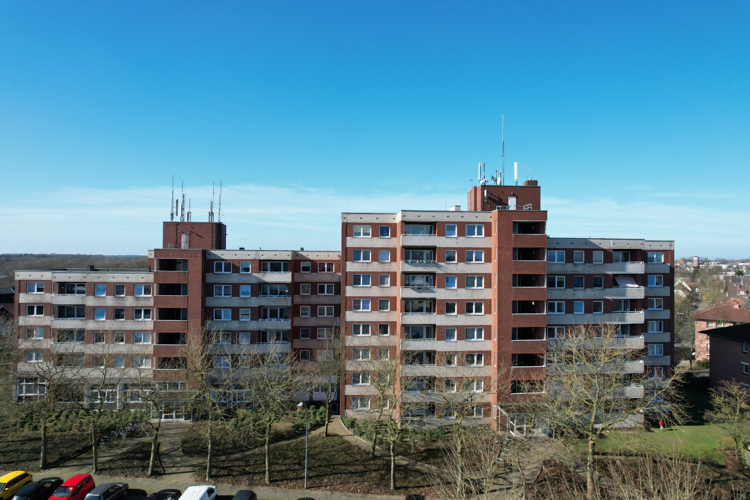 Ein mittelhoher roter Backsteinwohnungsbau mit Balkonen und Antennen auf dem Dach vor einem klaren blauen Himmel. Im Vordergrund sind blattlose B&auml;ume und geparkte Autos zu sehen.